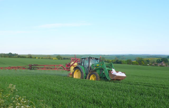 A tractor-mounted boom sprayer applying a treatment across a green cereal crop